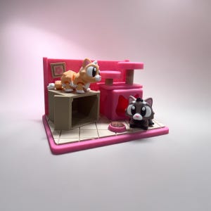 Puede incluir: Un juego de miniaturas con temática de gatos, con una base y paredes rosas. Cuenta con una figura de gato marrón y blanco sobre una caja marrón, una figura de gato gris y negro, un rascador y un cuenco de comida. El set tiene un diseño de dibujos animados.