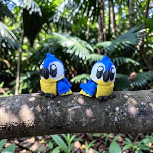 May include: Two small parrot figurines featuring bright blue wings, yellow chests, and white faces with large black eyes and black beaks. The figures are positioned side by side on a textured brown tree branch, showcasing a stylized design.