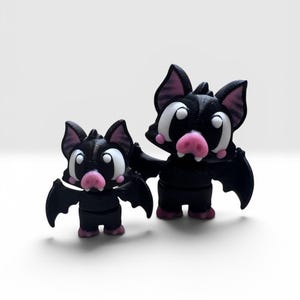 May include: Two black bat figurines of different sizes, each featuring large white eyes, a pink snout, pink inner ears, and small pink feet. The bats have pointed ears and outstretched wings, with a smooth, matte finish on their bodies.