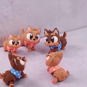 Op de afbeelding: Een verzameling van vijf kleine hondjes in cartoonstijl. De honden zijn voornamelijk bruin met witte en zwarte accenten, en dragen blauwe of roze polkadot kraagjes. Ze hebben grote, expressieve ogen en staan in verschillende houdingen.
