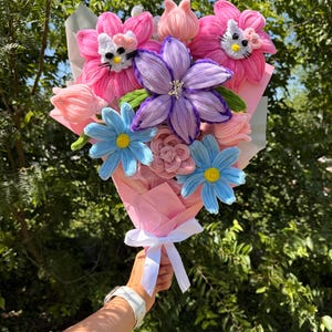 May include: A bouquet of crocheted flowers featuring two pink blossoms with Hello Kitty faces, a central purple lily, two light blue daisies, and several small pink buds. The arrangement is wrapped in pink paper and tied with a white ribbon.