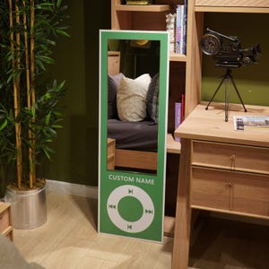 May include: A rectangular green-framed mirror with a white border. The bottom section features a white circle with play, forward, and rewind symbols. The text "CUSTOM NAME" is above the circle. The mirror reflects a bed with pillows.