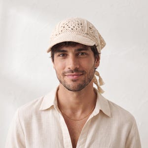 Puede incluir: Una gorra de ganchillo color crema con visera estructurada y una corona con patrón de cuadrados. La prenda está tejida con hilo de textura detallada e incluye dos lazos colgantes con pequeñas borlas en los extremos, atados en la parte posterior.