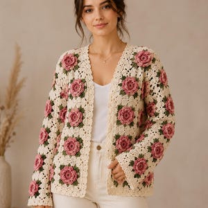 Könnte beinhalten: Cremefarbene Häkeljacke mit floralem Design aus rosa Rosen und grünen Blättern. Der Cardigan mit offenem Schnitt hat einen gewellten Rand und ist aus weichem, strukturiertem Garn gefertigt. Ein stilvolles, handgefertigtes Kleidungsstück.