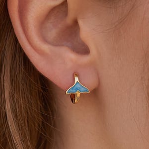 Könnte beinhalten: Ein goldener Creolen-Ohrring mit einem kleinen, blau emaillierten Walflossen-Anhänger. Der Anhänger ist in Form einer Fluke gestaltet und mittig am unteren Teil des Reifens befestigt. Der Ohrring sitzt im Ohrläppchen und zeigt ein poliertes Goldfinish.
