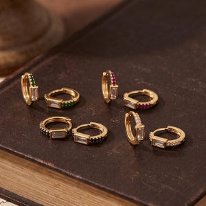 May include: A collection of gold hoop earrings featuring baguette-cut clear stones and small round accent stones in green, red, and black colors. Each earring is a small circular hoop with a hinged closure, showcasing a metallic gold finish.