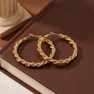 May include: A pair of gold-toned hoop earrings featuring a twisted rope-like texture. The earrings are circular in shape and have a polished metallic finish, showcasing a detailed braided design throughout the entire circumference of each hoop.