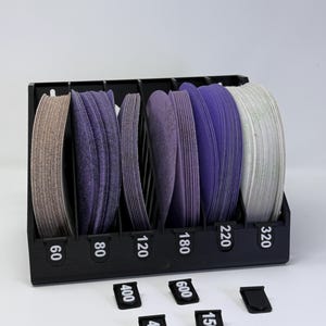 May include: A black organiser holding stacks of sanding discs in shades of tan, purple, and white. The organiser has labels with numbers such as 60, 80, 120, 180, 220, and 320. Additional labels with numbers 40, 150, 400, and 600 are also present.