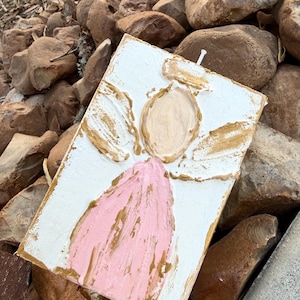 May include: A rectangular, textured, white painted canvas with an angel design. The angel features a pink robe, a beige face, and gold-colored accents on the wings and halo. A small white nail is visible at the top.
