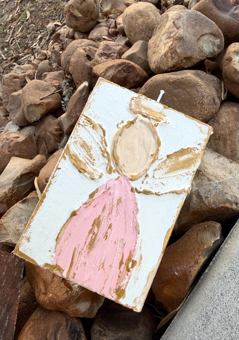 May include: A rectangular, textured, white painted canvas with an angel design. The angel features a pink robe, a beige face, and gold-colored accents on the wings and halo. A small white nail is visible at the top.