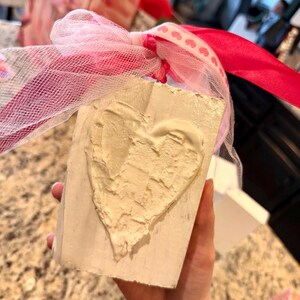 May include: A rectangular, off-white block with a textured, cream-colored heart design. The block is tied with a pink ribbon and white mesh. The background includes other gift boxes with pink and white accents.