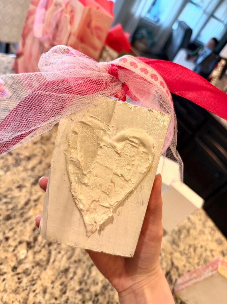 May include: A rectangular, off-white block with a textured, cream-colored heart design. The block is tied with a pink ribbon and white mesh. The background includes other gift boxes with pink and white accents.