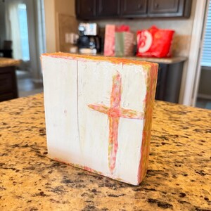May include: A white wooden block with a hand-painted cross in shades of pink and red. The edges of the block are painted with pink and yellow hues. The artwork is displayed on a speckled countertop.