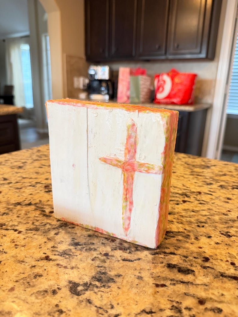 May include: A white wooden block with a hand-painted cross in shades of pink and red. The edges of the block are painted with pink and yellow hues. The artwork is displayed on a speckled countertop.