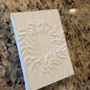 May include: A white, rectangular wooden plaque with a raised, textured wreath design. The wreath is composed of stylized leaves. The plaque is painted white and has a rustic, handmade appearance, suitable for home decor.