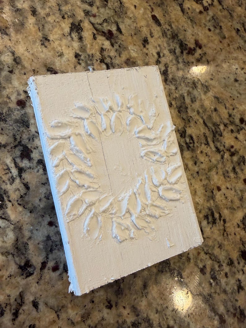 May include: A white, rectangular wooden plaque with a raised, textured wreath design. The wreath is composed of stylized leaves. The plaque is painted white and has a rustic, handmade appearance, suitable for home decor.