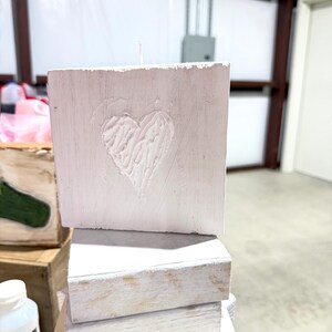 May include: A stack of three white wooden blocks, with the top block featuring a carved heart design. The blocks have a rustic, distressed appearance, suggesting a handmade or vintage aesthetic. The candle is white.