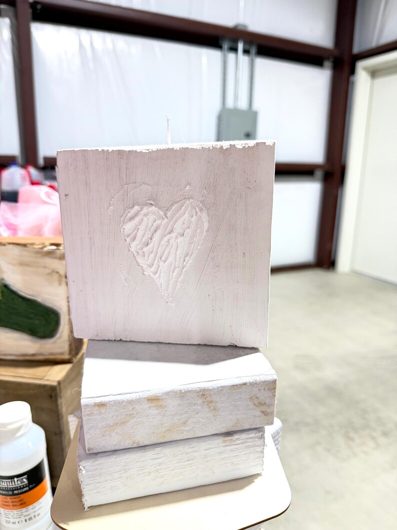 May include: A stack of three white wooden blocks, with the top block featuring a carved heart design. The blocks have a rustic, distressed appearance, suggesting a handmade or vintage aesthetic. The candle is white.