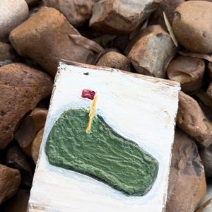 May include: A small, rectangular, hand-painted wooden sign featuring a green golf course with a red flag on a yellow pole. The sign has a white background and a brown border. The sign is resting on a bed of brown rocks.