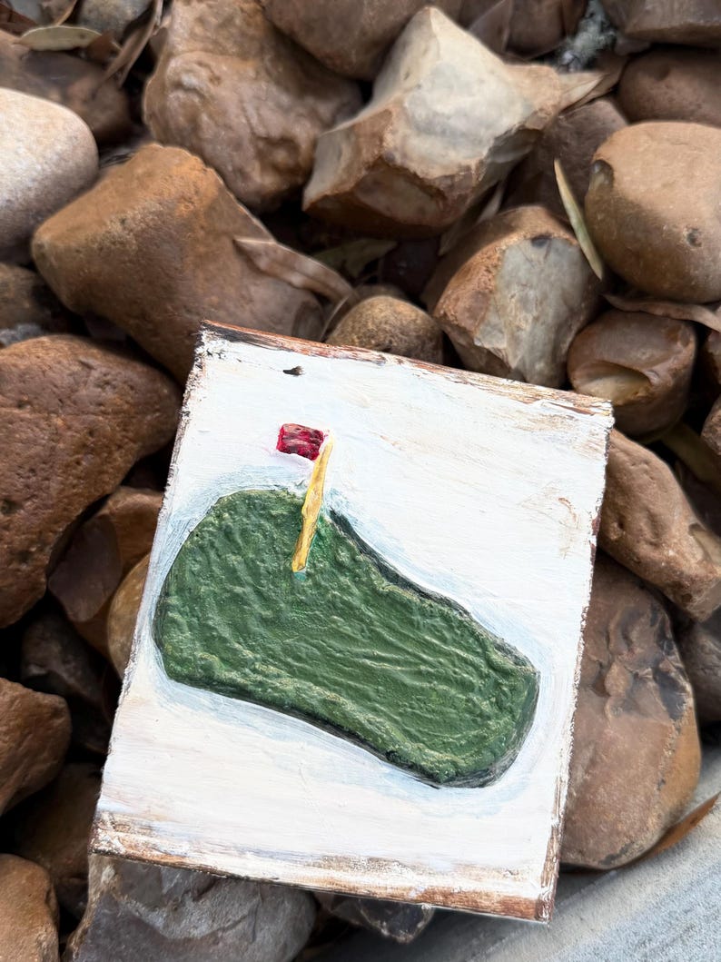 May include: A small, rectangular, hand-painted wooden sign featuring a green golf course with a red flag on a yellow pole. The sign has a white background and a brown border. The sign is resting on a bed of brown rocks.