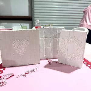 May include: Three white, textured wooden blocks with carved designs. One block features a heart, another a cross, and the third a wreath. The blocks are arranged on a pink surface with red heart-shaped cutouts.