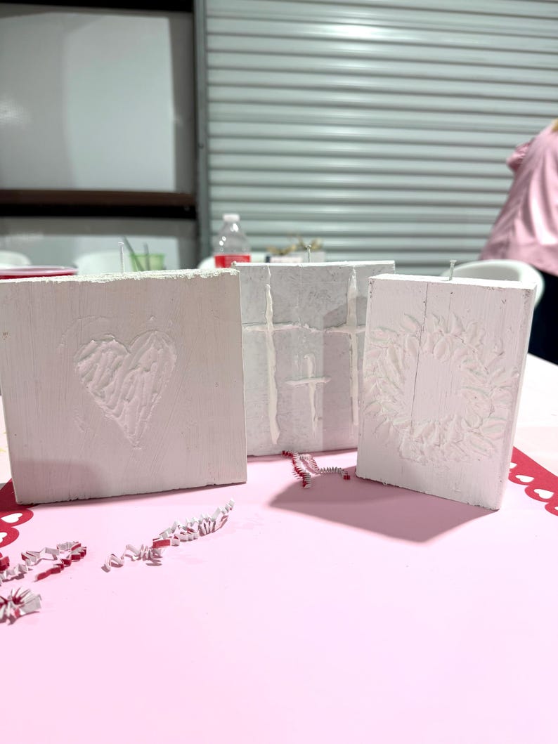 May include: Three white, textured wooden blocks with carved designs. One block features a heart, another a cross, and the third a wreath. The blocks are arranged on a pink surface with red heart-shaped cutouts.
