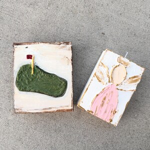 May include: Two small, rectangular, hand-painted artworks. One depicts a green golf course with a red flag, the other an angel in pink and gold. Both are on white backgrounds with textured paint.