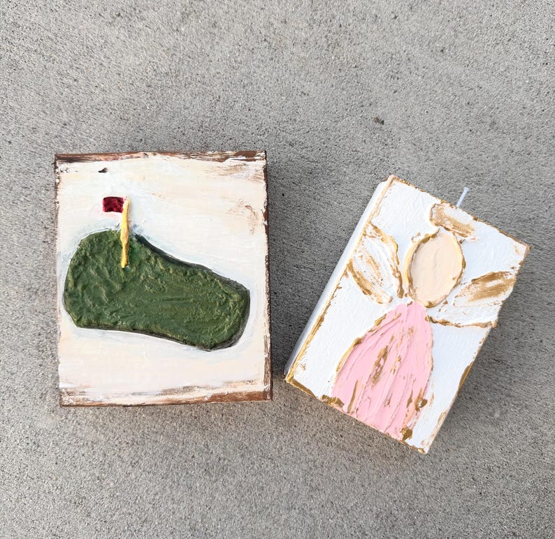 May include: Two small, rectangular, hand-painted artworks. One depicts a green golf course with a red flag, the other an angel in pink and gold. Both are on white backgrounds with textured paint.