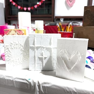 May include: Three white textured art canvases with embossed designs: a floral pattern, a cross, and a heart. The canvases are displayed on a white surface, suggesting a craft or art project. Valentine's Day decorations are visible in the background.