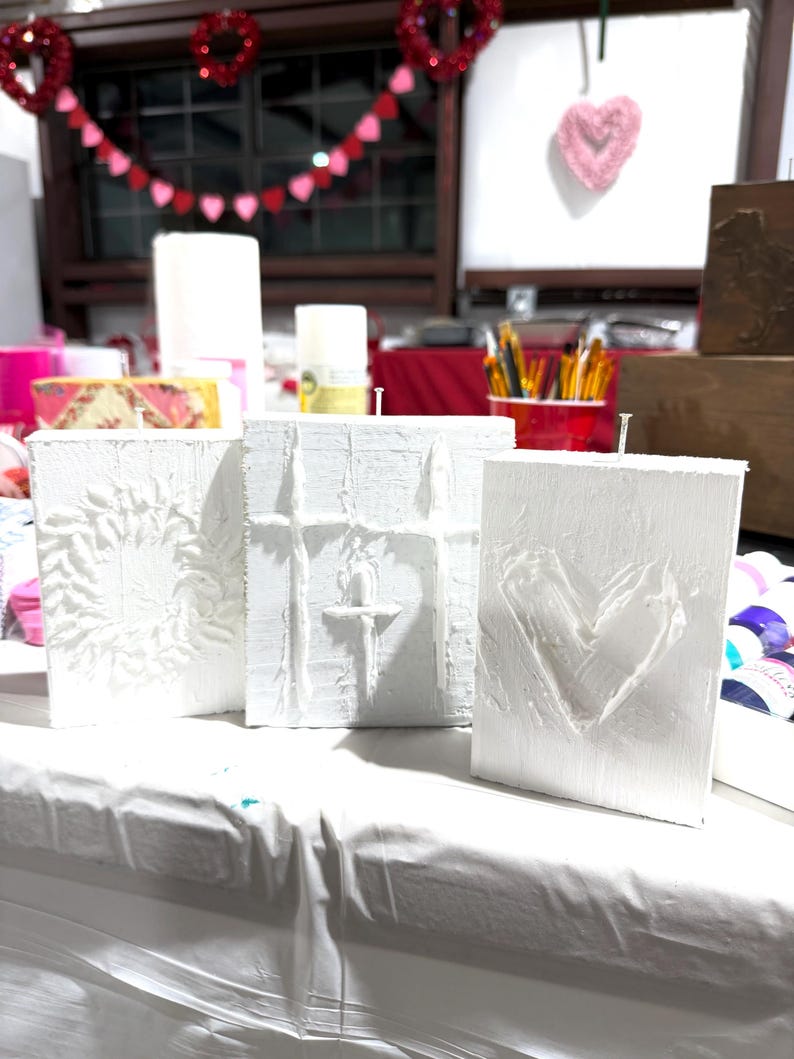 May include: Three white textured art canvases with embossed designs: a floral pattern, a cross, and a heart. The canvases are displayed on a white surface, suggesting a craft or art project. Valentine's Day decorations are visible in the background.