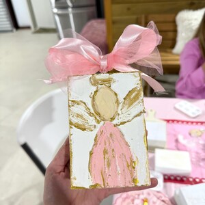 May include: A hand-painted angel on a white wooden block, featuring a pink dress and gold accents. The angel has a gold halo and wings. A pink ribbon and tulle bow adorn the top. The artwork is held in a hand.