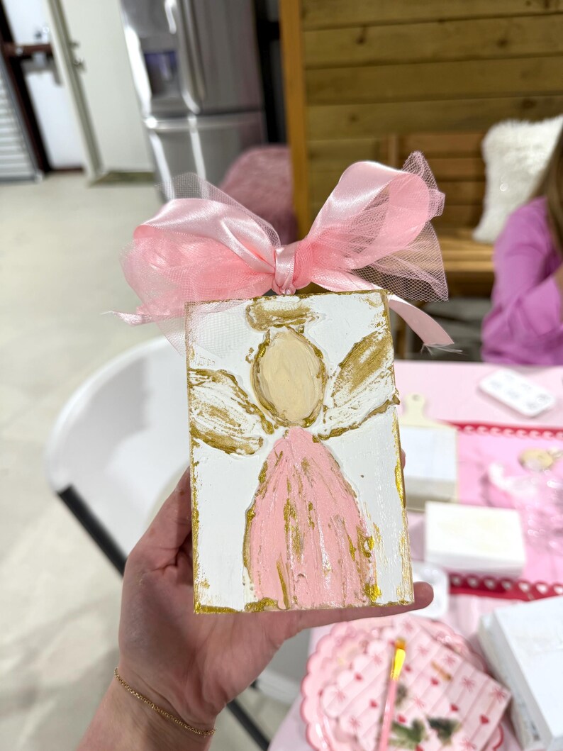 May include: A hand-painted angel on a white wooden block, featuring a pink dress and gold accents. The angel has a gold halo and wings. A pink ribbon and tulle bow adorn the top. The artwork is held in a hand.