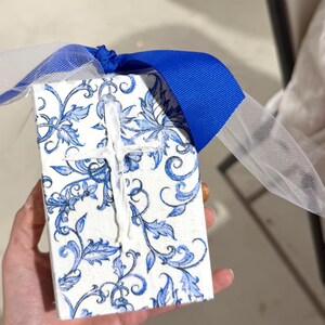 May include: A small, rectangular gift box with a white and blue floral pattern. A white cross is affixed to the top. The box is tied with a blue ribbon and white tulle.