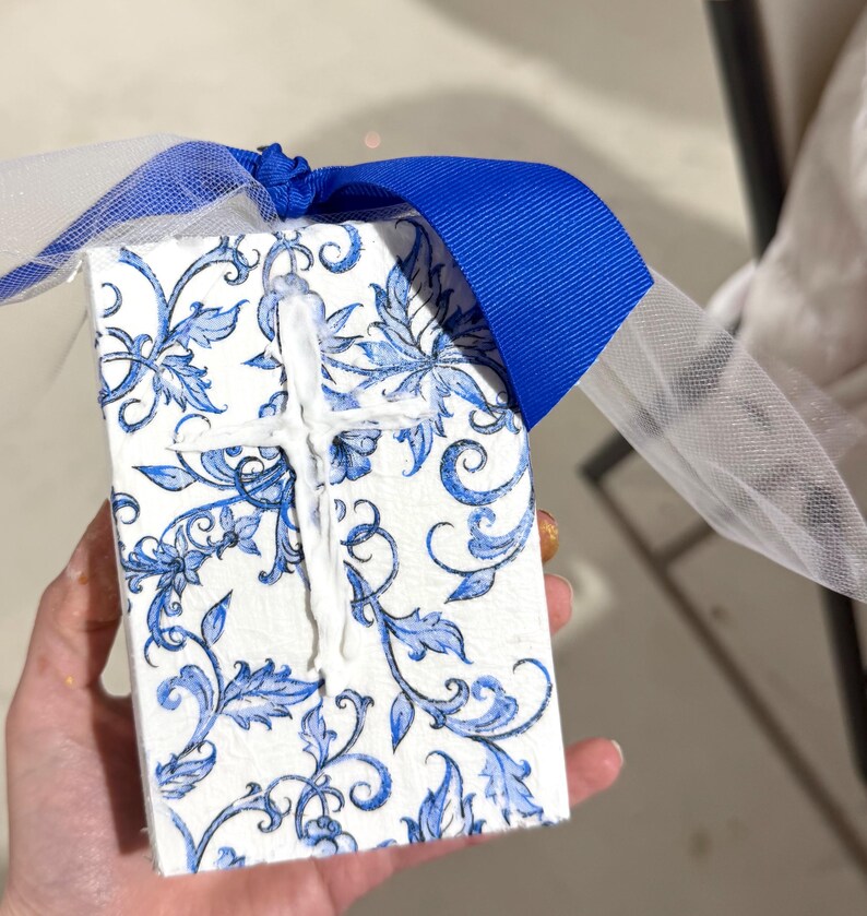 May include: A small, rectangular gift box with a white and blue floral pattern. A white cross is affixed to the top. The box is tied with a blue ribbon and white tulle.