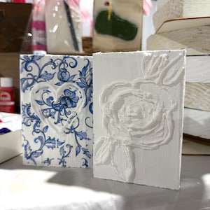 May include: Two decorative rectangular blocks. One features a blue and white floral pattern with a heart design. The other is white with a raised rose design. Both are displayed on a white surface.