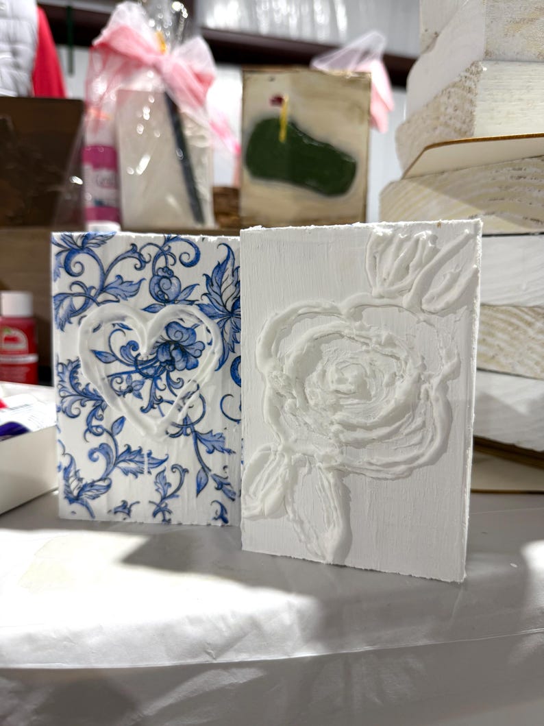 May include: Two decorative rectangular blocks. One features a blue and white floral pattern with a heart design. The other is white with a raised rose design. Both are displayed on a white surface.