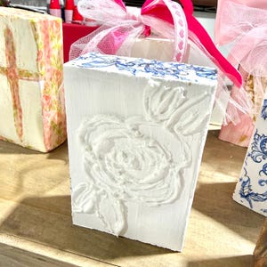 May include: A white rectangular block with a raised floral design and blue accents. A pink ribbon and sheer white fabric are tied on top. Other decorative blocks with floral and patterned designs are in the background.
