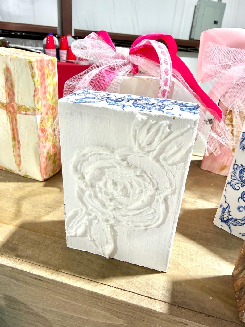 May include: A white rectangular block with a raised floral design and blue accents. A pink ribbon and sheer white fabric are tied on top. Other decorative blocks with floral and patterned designs are in the background.