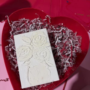 May include: A white rectangular canvas with a textured floral design of roses in a vase. The canvas is placed in a red heart-shaped tray filled with red and white shredded paper, set on a pink surface.