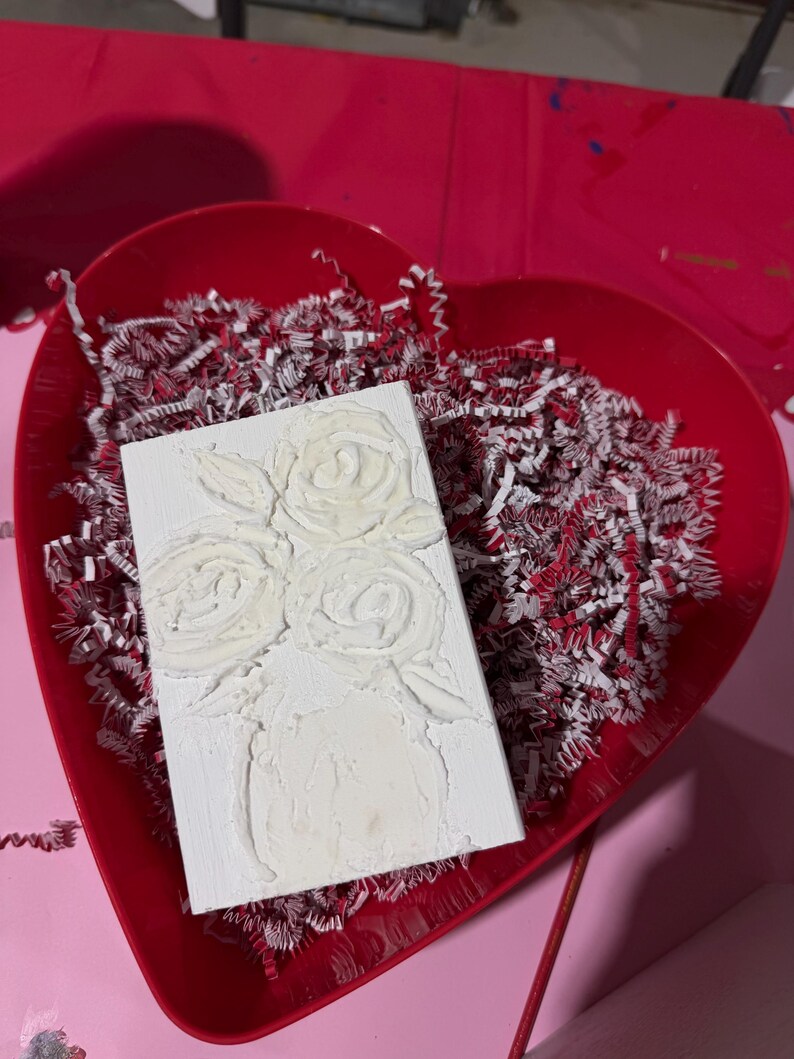 May include: A white rectangular canvas with a textured floral design of roses in a vase. The canvas is placed in a red heart-shaped tray filled with red and white shredded paper, set on a pink surface.