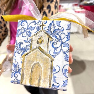May include: A small, square, hand-painted ornament featuring a church design. The church is painted in gold with a cross on top, set against a white background with blue floral patterns. A gold and white ribbon is attached for hanging.