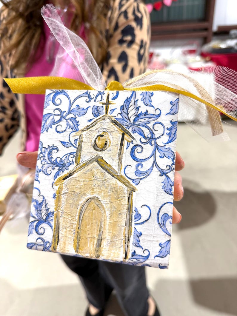 May include: A small, square, hand-painted ornament featuring a church design. The church is painted in gold with a cross on top, set against a white background with blue floral patterns. A gold and white ribbon is attached for hanging.