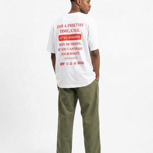 Könnte beinhalten: Ein weißes T-Shirt mit rotem Text, der lautet: "FOR A PHILTHY TIME, CALL 0735-696969 WHY BE MOODY, IF YOU CAN SHAKE YOUR BOOTY." Die Person trägt eine olivgrüne Hose und schwarze Schuhe.