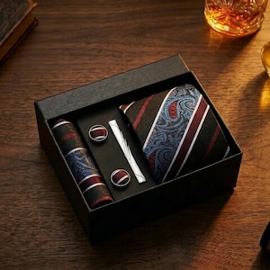 May include: A black gift box containing a necktie, a tie clip, and two cufflinks. The necktie features a pattern of blue paisley, black stripes, and thin red and white lines. The matching cufflinks and the tie clip have a silver-tone finish.