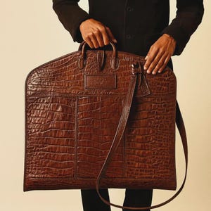 May include: A brown leather garment bag with a crocodile texture. The bag has a top handle, a shoulder strap, and a rectangular label. The bag is held by a person wearing a black jacket.