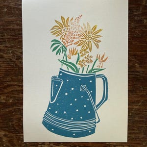 May include: A print of a blue watering can filled with flowers. The watering can has white polka dots and a handle. The flowers are yellow, orange, and green. The print is on a cream-colored background.