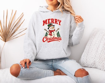 May include: A light gray hooded sweatshirt featuring a graphic of a snowman wearing a black top hat and red scarf, flanked by two green pine trees. The text Merry Christmas is printed in red above and below the snowman illustration.
