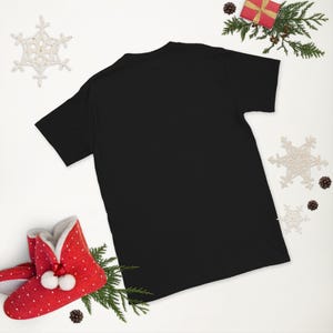 Puede incluir: Una camiseta negra, vista desde atrás, es el foco central de esta imagen. La camiseta está rodeada de decoraciones navideñas, incluyendo una caja de regalo roja, ramitas de pino, copos de nieve y un par de zapatillas rojas con lunares blancos.