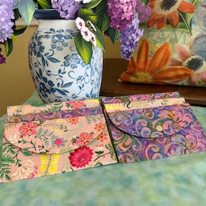 May include: Two fabric pouches with floral patterns in pink and purple, featuring snap closures. A blue and white floral vase holds paper flowers. A floral pillow and a green and blue surface complete the scene.