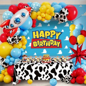 May include: A festive birthday party scene with a blue backdrop featuring white clouds and the words "Happy Birthday." Balloons in red, yellow, blue, and cow print decorate the scene, along with a rocket ship balloon and a table with a cow print tablecloth.